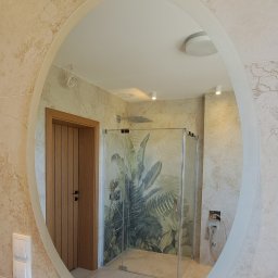 WB-Qube Waldemar Bednarczyk - Owalne lustro w łazience z beżowymi płytkami imitującymi marmur, odbijające kabinę prysznicową z motywem roślinnym i drewniane drzwi. Nowoczesna armatura.