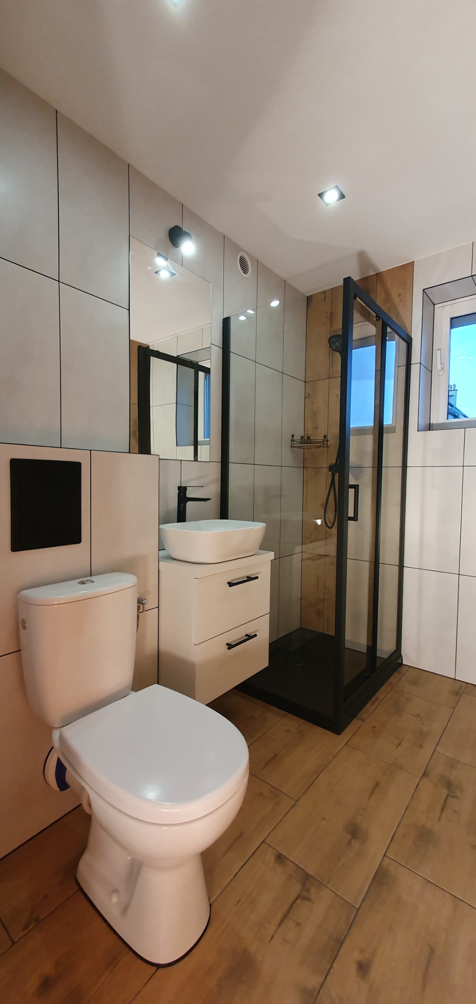 Nowoczesna łazienka z kabiną prysznicową typu walk-in z czarnymi ramami, umywalką nablatową na szafce z szufladami i podwieszaną toaletą, wykończona płytkami w odcieniach bieli i beżu...