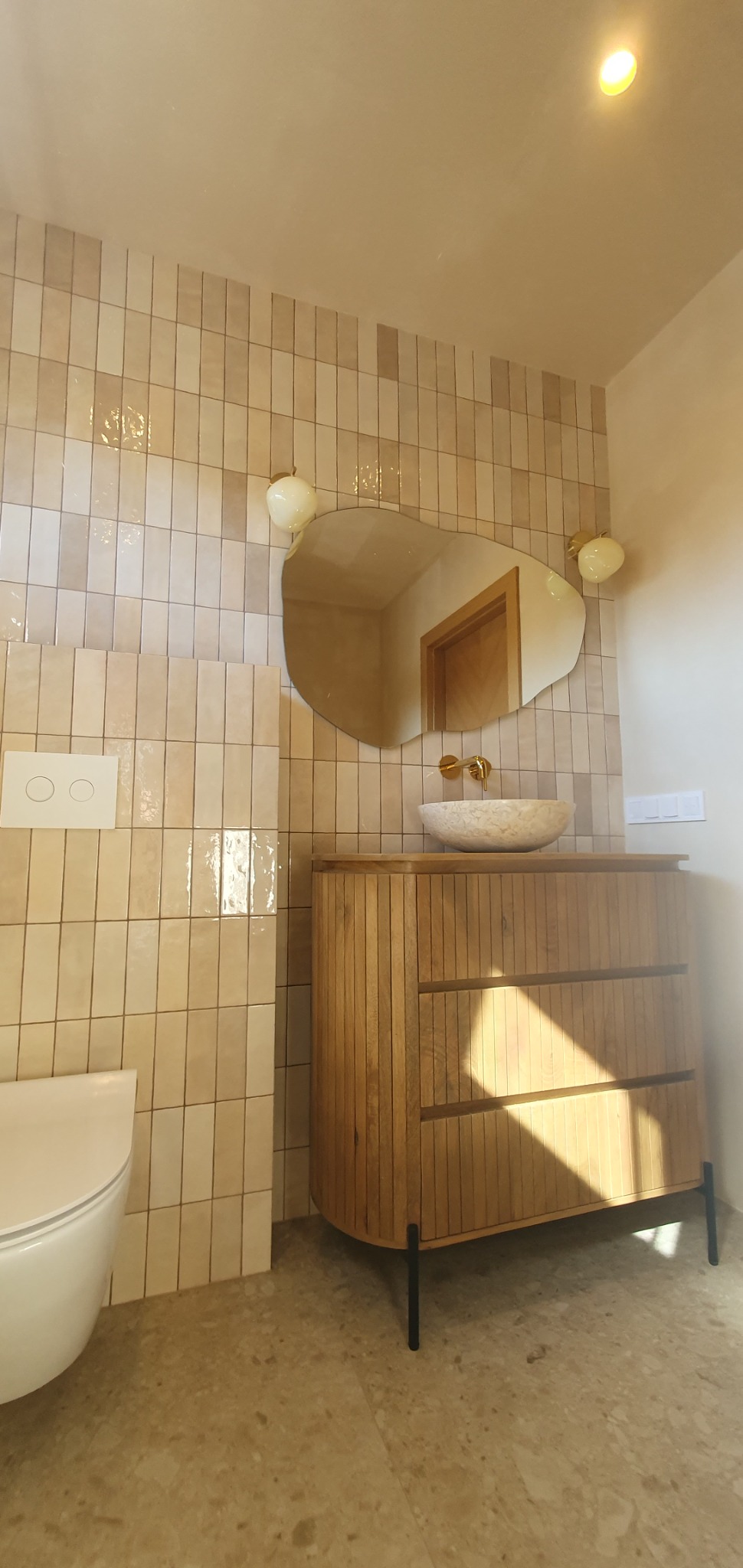 Wykończona łazienka z beżowymi, pionowo ułożonymi płytkami, asymetrycznym lustrem, kamienną umywalką nablatową, drewnianą szafką z trzema szufladami i wiszącą miską WC.