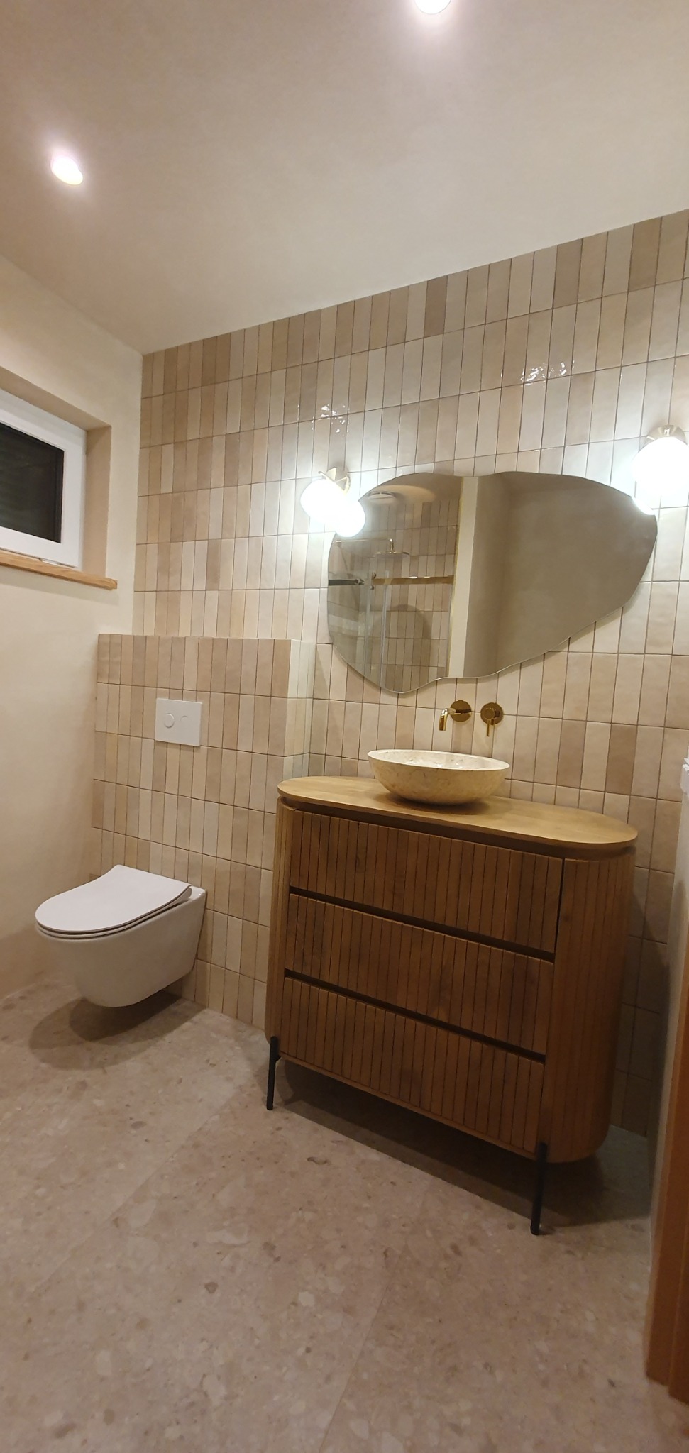 Ukończona łazienka w beżowych odcieniach z umywalką nablatową, drewnianą szafką z ryflowanymi frontami, asymetrycznym lustrem i podwieszaną toaletą.