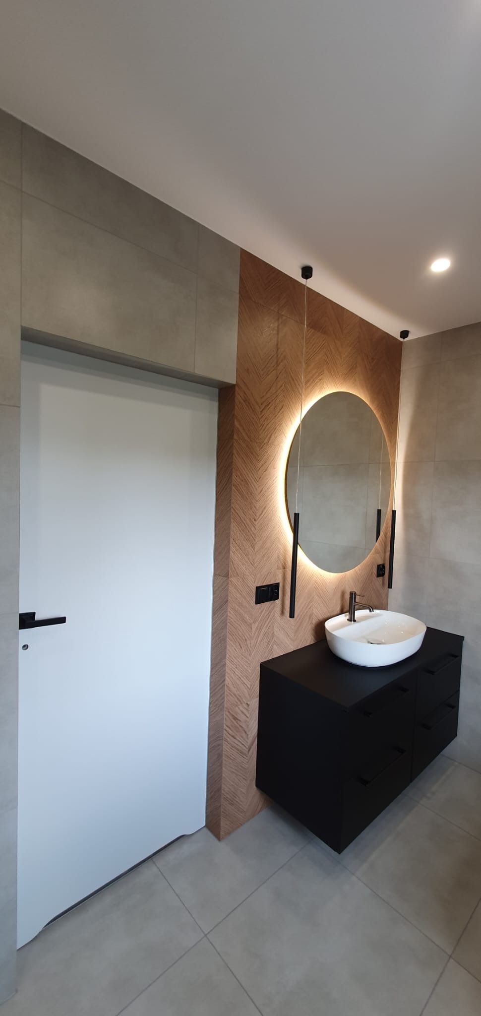 Nowoczesna łazienka z okrągłym lustrem podświetlanym LED, czarną szafką pod umywalką i drewnianą ścianą w jodełkę. Minimalistyczny design, szare płytki podłogowe.