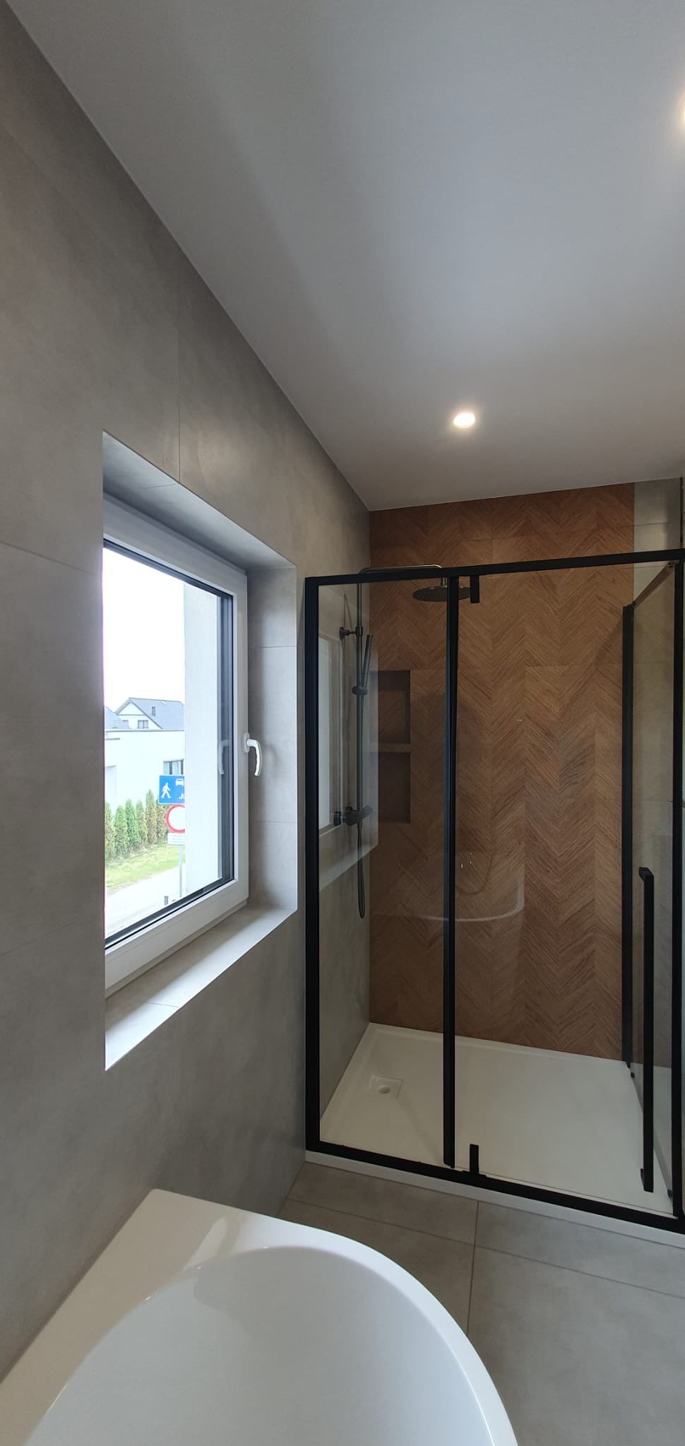 Nowoczesna łazienka z kabiną typu walk-in z czarnymi profilami i drewnopodobną ścianą w jodełkę, widok zza białej wanny, okno z widokiem na zewnątrz.