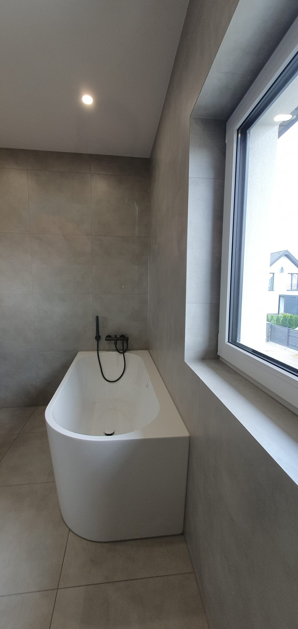 Nowoczesna łazienka z wolnostojącą białą wanną i czarną armaturą na tle szarych płytek i okna z widokiem na dom.