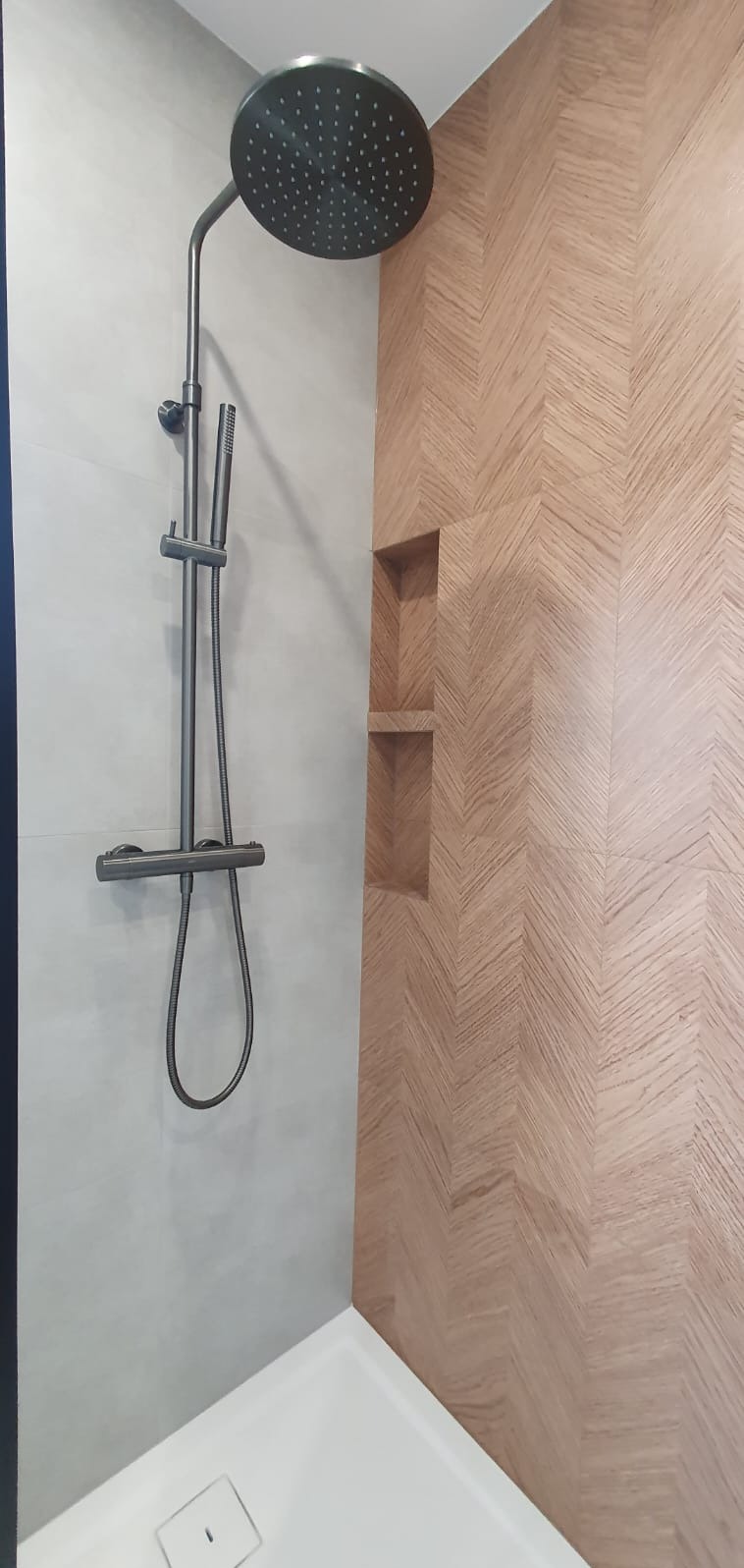 Nowoczesna kabina prysznicowa z deszczownicą i półkami w ścianie wyłożonej płytkami imitującymi drewno ułożone w jodełkę. Szara ściana i biały brodzik.