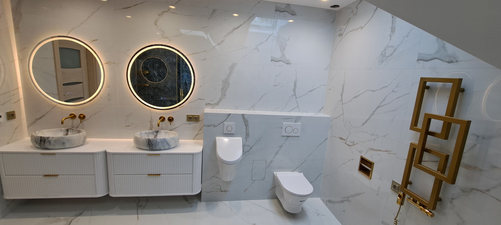 Elegancka łazienka w bieli i marmurze z okrągłymi lustrami podświetlanymi LED, umywalkami z marmuru i złotym grzejnikiem drabinkowym.
