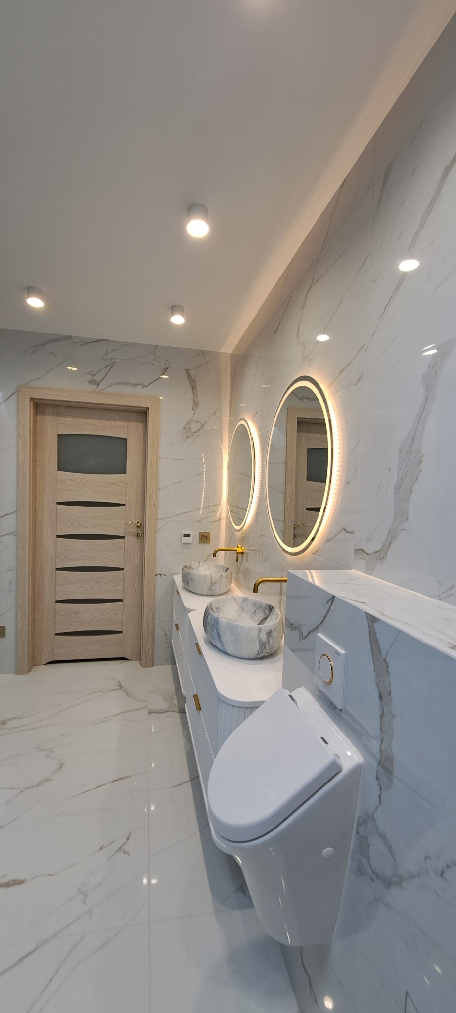 Elegancka łazienka z marmurowymi płytkami, podwójną umywalką w stylu vessel, okrągłymi lustrami z podświetleniem LED i wiszącą toaletą.