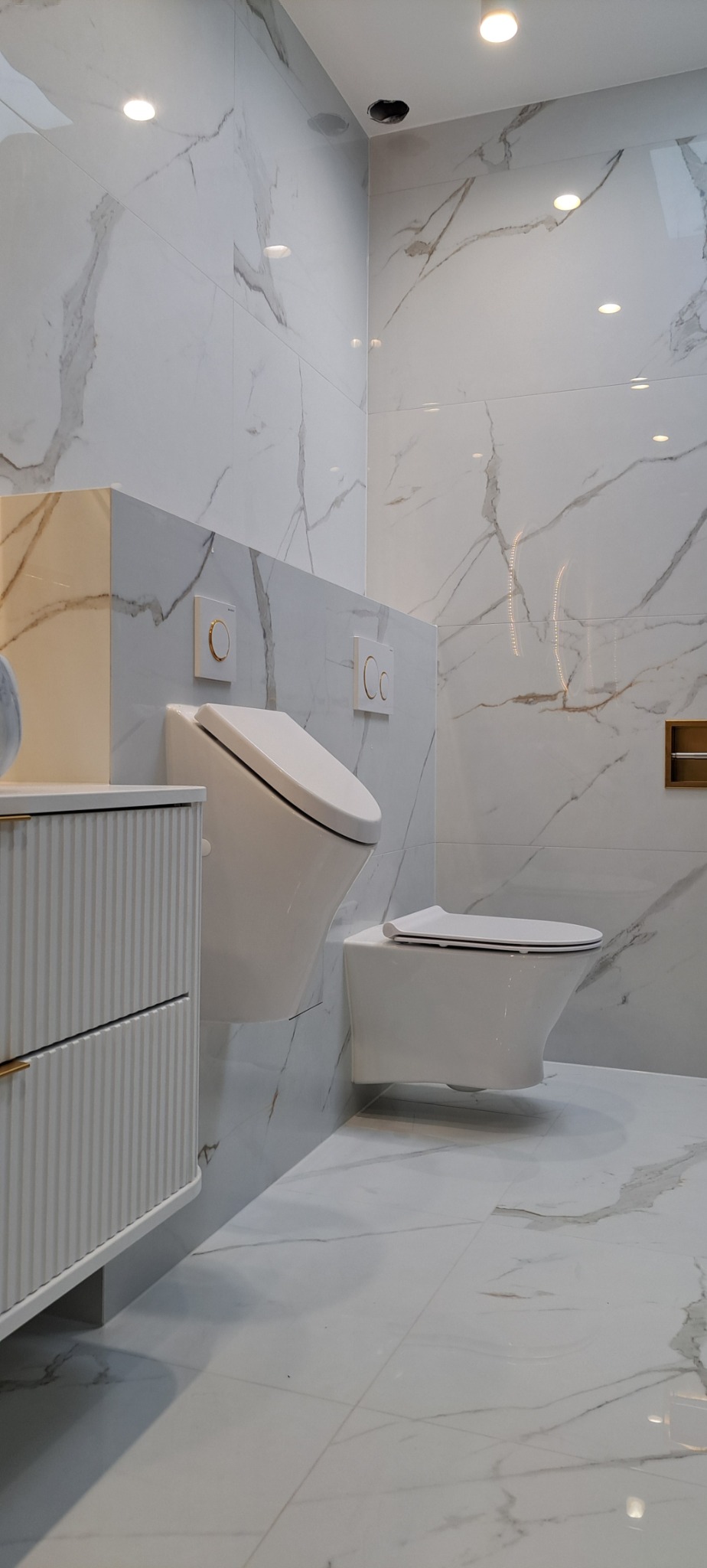 Elegancka łazienka z marmurowymi płytkami, podwieszanymi toaletami i szafką z ryflowanymi frontami. Oświetlenie punktowe w suficie.