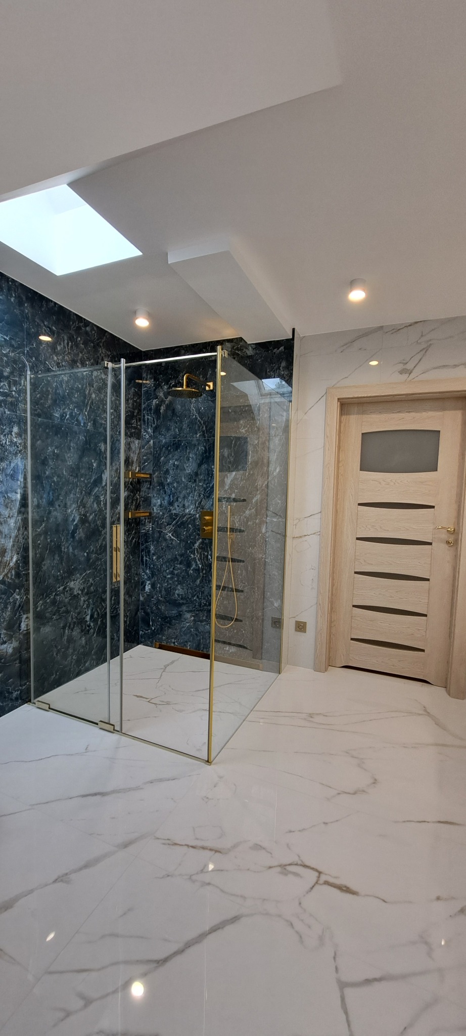 Nowoczesna łazienka po remoncie: kabina prysznicowa ze złotymi detalami, marmurowe płytki na ścianach i podłodze, oświetlenie sufitowe.
