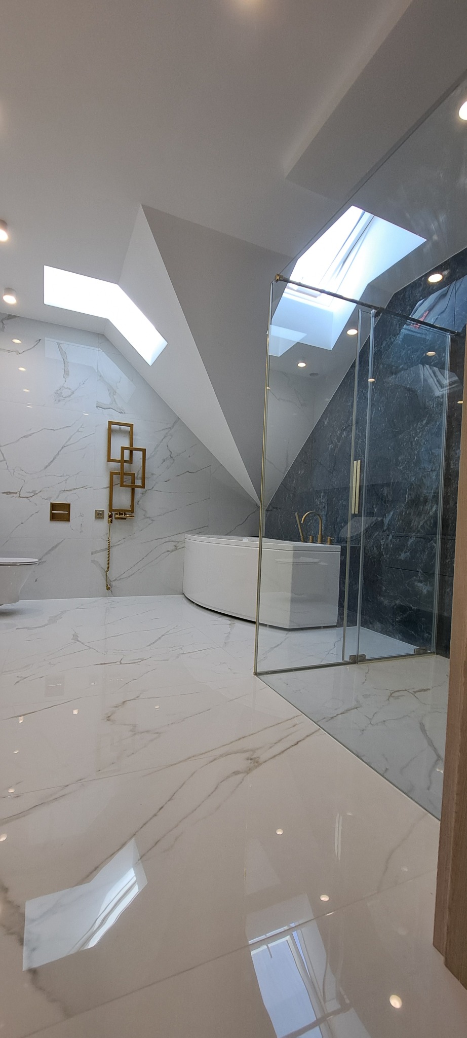 Elegancka łazienka na poddaszu z biało-szarymi marmurowymi płytkami, białą wanną i złotymi akcentami. Widoczne okno dachowe i szklana kabina prysznicowa.