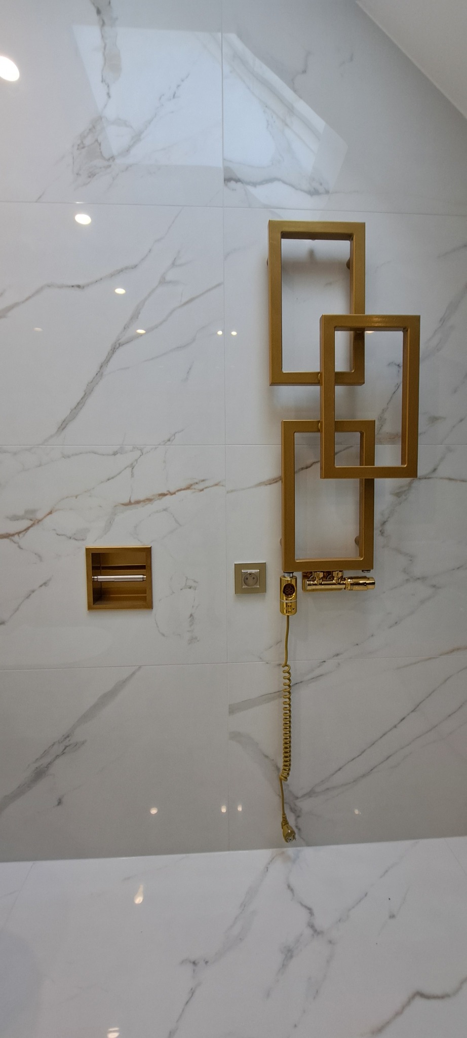 Białe płytki imitujące marmur z złotymi akcentami: grzejnik drabinkowy, uchwyt na papier toaletowy i gniazdko. Nowoczesny design łazienki w eleganckim wydaniu.