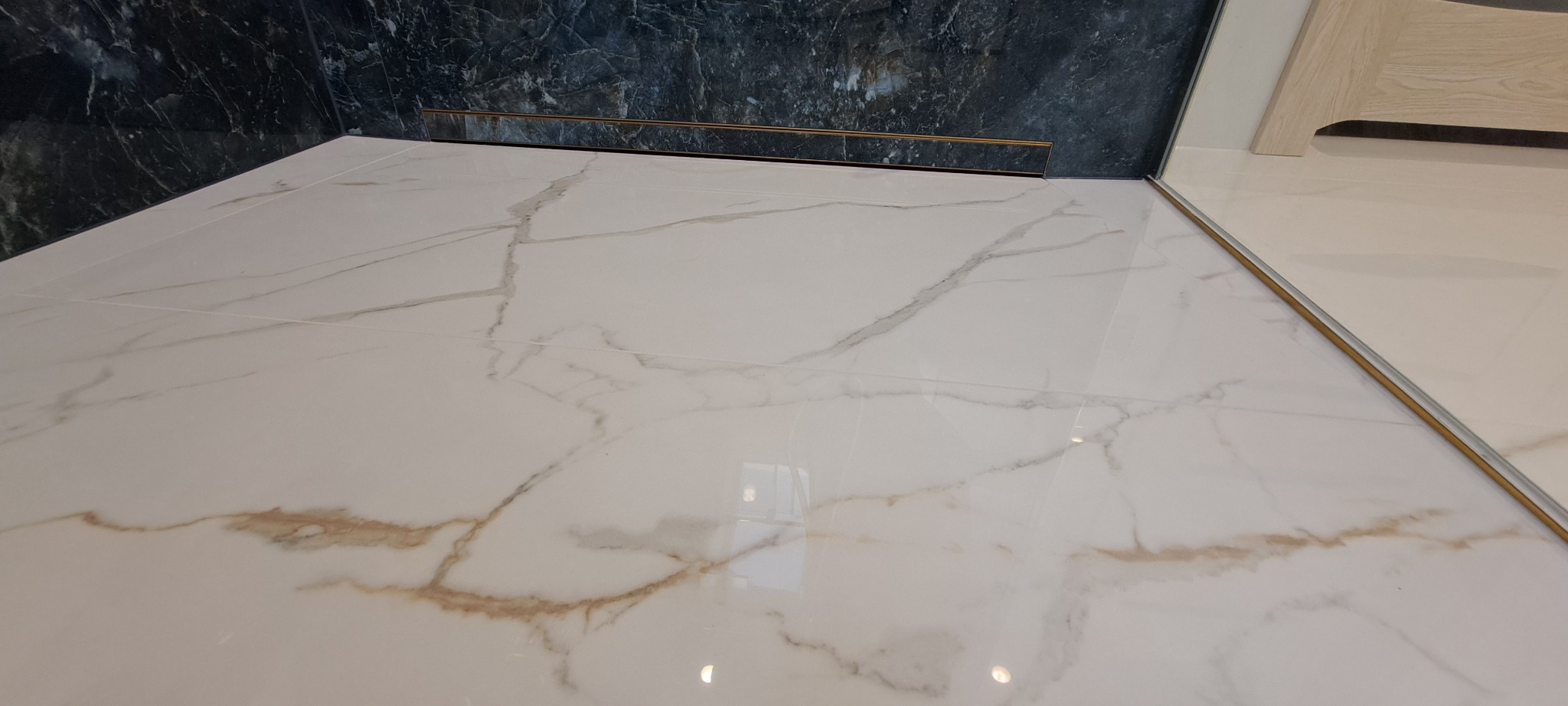 Błyszczące, marmurowe płytki podłogowe w łazience z odpływem liniowym i fragmentem ciemnej ściany imitującej kamień. Widoczny narożnik zabudowy.