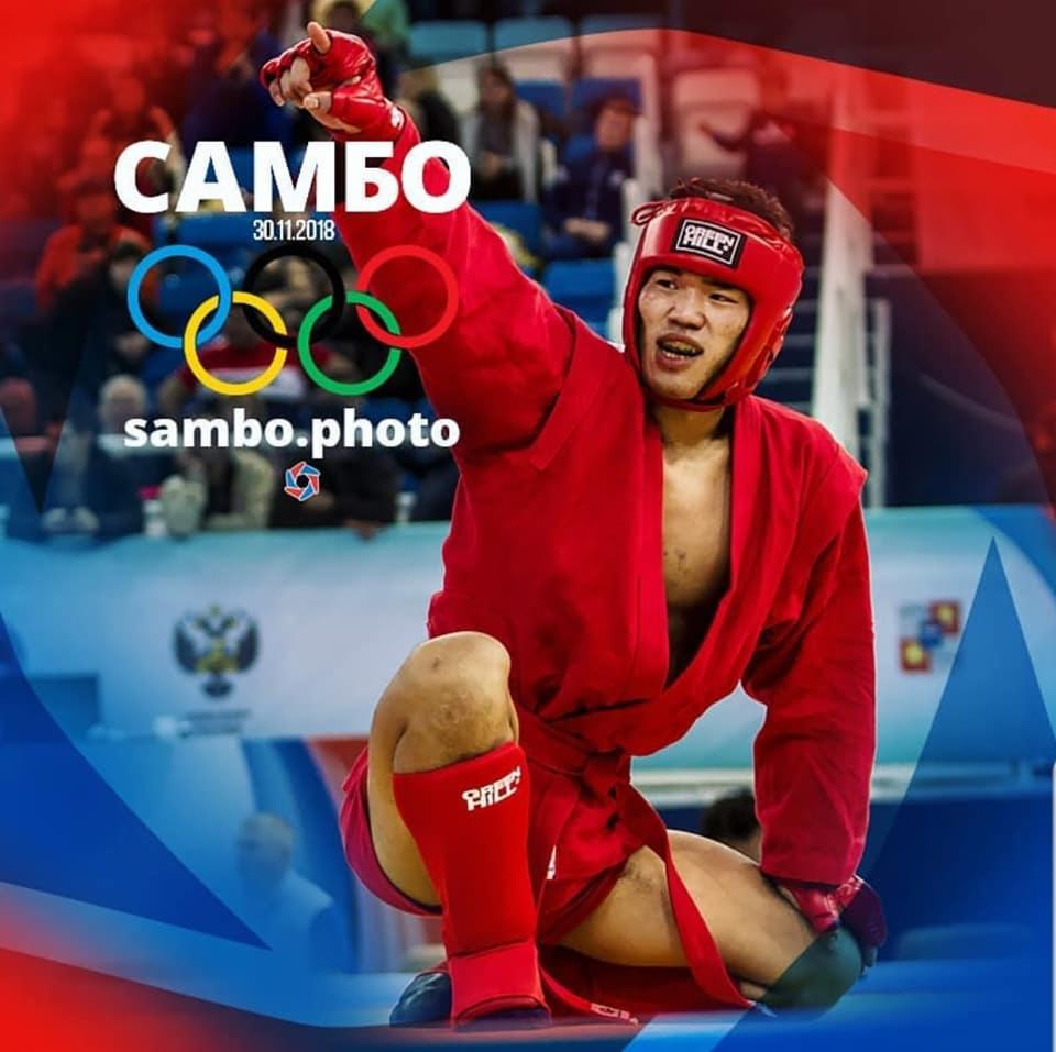 Zawodowy sportowiec w czerwonym stroju sambo, unosi rękę w geście zwycięstwa na tle publiczności.