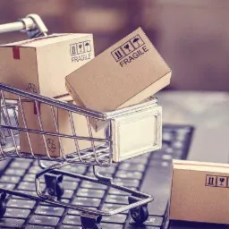 Miniaturowy wózek sklepowy wypełniony kartonowymi pudełkami z napisem 'FRAGILE' na klawiaturze laptopa, stonowana kolorystyka.
