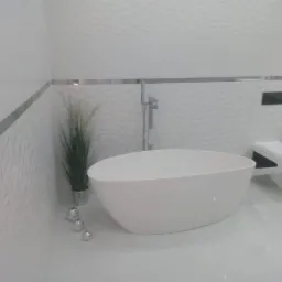 Nowoczesna, minimalistyczna łazienka z wolnostojącą białą wanną, trójwymiarowymi białymi płytkami ściennymi, srebrnymi listwami dekoracyjnymi, wiszącą toaletą i rośliną w srebrnej doniczce.