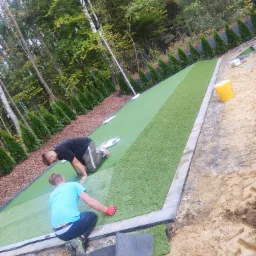 Dwaj mężczyźni układają sztuczną trawę na przygotowanym podłożu w ogrodzie, widoczne obrzeża z kostki, w tle drzewa i tuje.