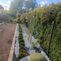 Pracownik w zielonej kamizelce układa białe kamienie na czarnej geowłókninie wzdłuż nowo posadzonych krzewów i drzewek, widoczna obrzeże z cegły i zielony wąż ogrodowy w tle.