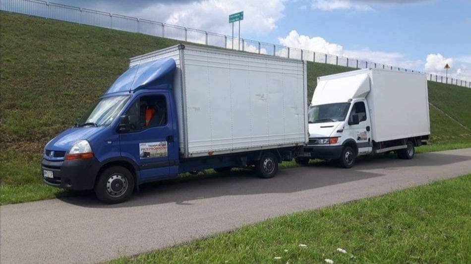 Dwa busy dostawcze, niebieski Renault Master i biały Iveco Daily, zaparkowane na poboczu drogi z zielonym wzgórzem w tle, sugerujące transport lub przeprowadzkę.