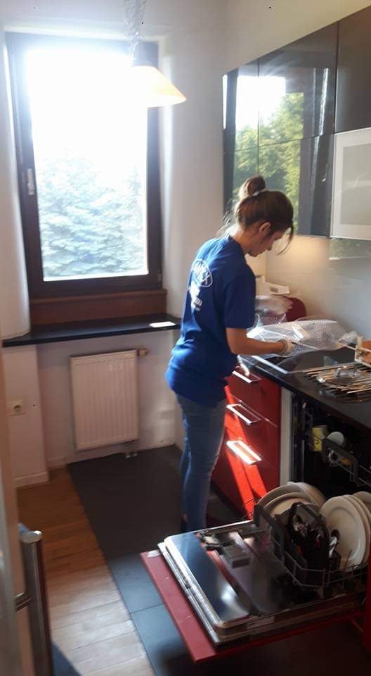 Kobieta w niebieskiej koszulce z logo firmy sprzątającej, w rękawicach, myje zlewozmywak w kuchni z czerwonymi szafkami, przy otwartych drzwiach zmywarki.