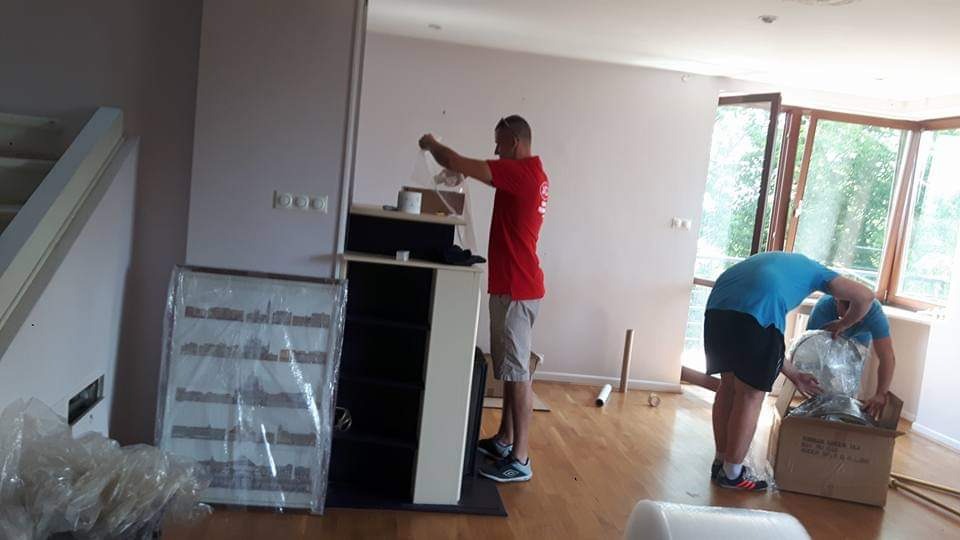 Dwóch mężczyzn pakuje i zabezpiecza meble folią stretch podczas przeprowadzki w jasnym, przestronnym pokoju z parkietem.