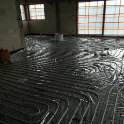 Niedokończona instalacja ogrzewania podłogowego w surowym stanie, rury ułożone na siatce, okna zabezpieczone folią ochronną, widoczny bęben z rurą i karton na podłodze.