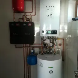 Zestaw grzewczy Buderus z zasobnikiem, kotłem i naczyniami przeponowymi, rurami miedzianymi, zamontowany na ścianie z płytek ceramicznych w pomieszczeniu technicznym.