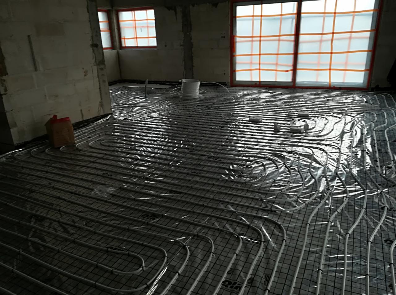 Niedokończona instalacja ogrzewania podłogowego w surowym stanie, rury ułożone na siatce, okna zabezpieczone folią ochronną, widoczny bęben z rurą i karton na podłodze.