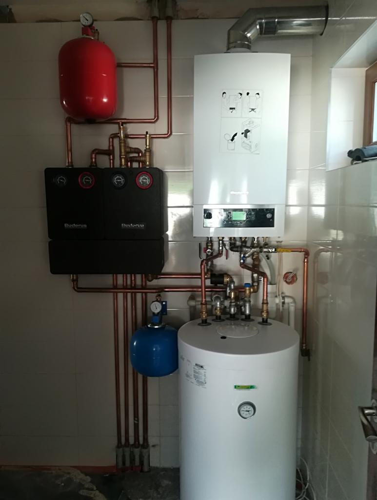 Zestaw grzewczy Buderus z zasobnikiem, kotłem i naczyniami przeponowymi, rurami miedzianymi, zamontowany na ścianie z płytek ceramicznych w pomieszczeniu technicznym.
