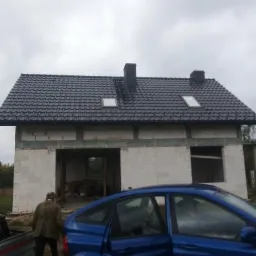 Dom w trakcie budowy z nowym, czarnym dachem z dachówki ceramicznej, dwoma oknami dachowymi i dwoma kominami, na pierwszym planie zaparkowany niebieski samochód z otwartymi drzwiami.