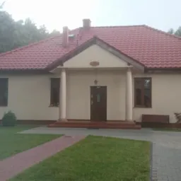Budynek parterowy z czerwonym dachem, kolumnami przy wejściu i zadbanym trawnikiem, otoczony drzewami.