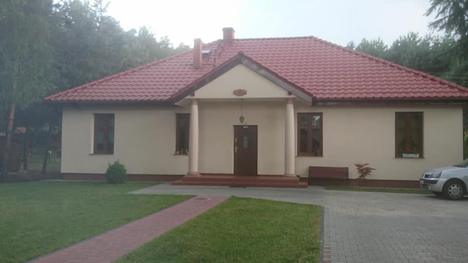 Budynek parterowy z czerwonym dachem, kolumnami przy wejściu i zadbanym trawnikiem, otoczony drzewami.
