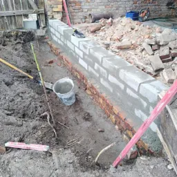 Wznoszona ściana fundamentowa z cegły i bloczków betonowych na zaprawie, widoczne narzędzia murarskie, poziomica i wiadro, otoczenie placu budowy z ziemią i gruzem.