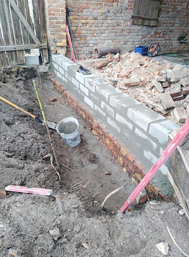 Wznoszona ściana fundamentowa z cegły i bloczków betonowych na zaprawie, widoczne narzędzia murarskie, poziomica i wiadro, otoczenie placu budowy z ziemią i gruzem.