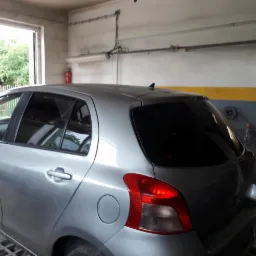 Srebrny samochód Toyota Yaris z przyciemnionymi szybami bocznymi i tylną, zaparkowany w garażu z widocznym oknem i wyposażeniem warsztatowym.