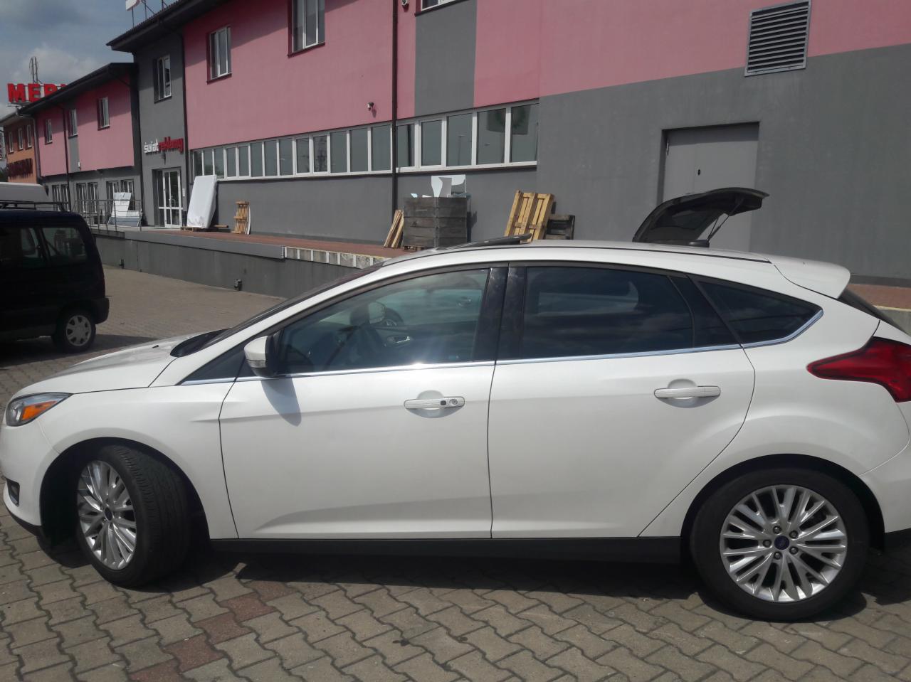 Biały Ford Focus hatchback z otwartą klapą bagażnika, zaparkowany na brukowanym parkingu przed budynkiem w odcieniach szarości i różu.