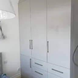 Biała, wysoka szafa z połyskiem, z trzema pionowymi segmentami drzwi i sześcioma szufladami z podłużnymi, metalowymi uchwytami, widoczna w sypialni z fragmentem łóżka i lampą.