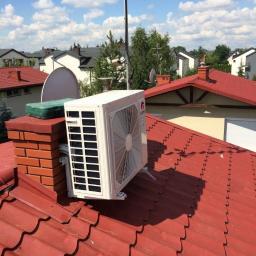 Klimatyzacja Pruszków - Biała jednostka zewnętrzna klimatyzatora marki Gree zamontowana na dachu z czerwonej dachówki obok komina z cegły, na tle błękitnego nieba z białymi chmurami i zabudowy jednorodzinnej.