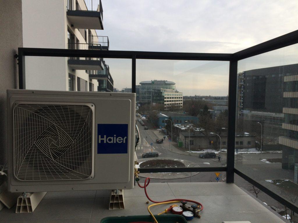 Jednostka zewnętrzna Haier na balkonie nowoczesnego apartamentowca, widoczne narzędzia serwisowe i panorama miasta w tle.