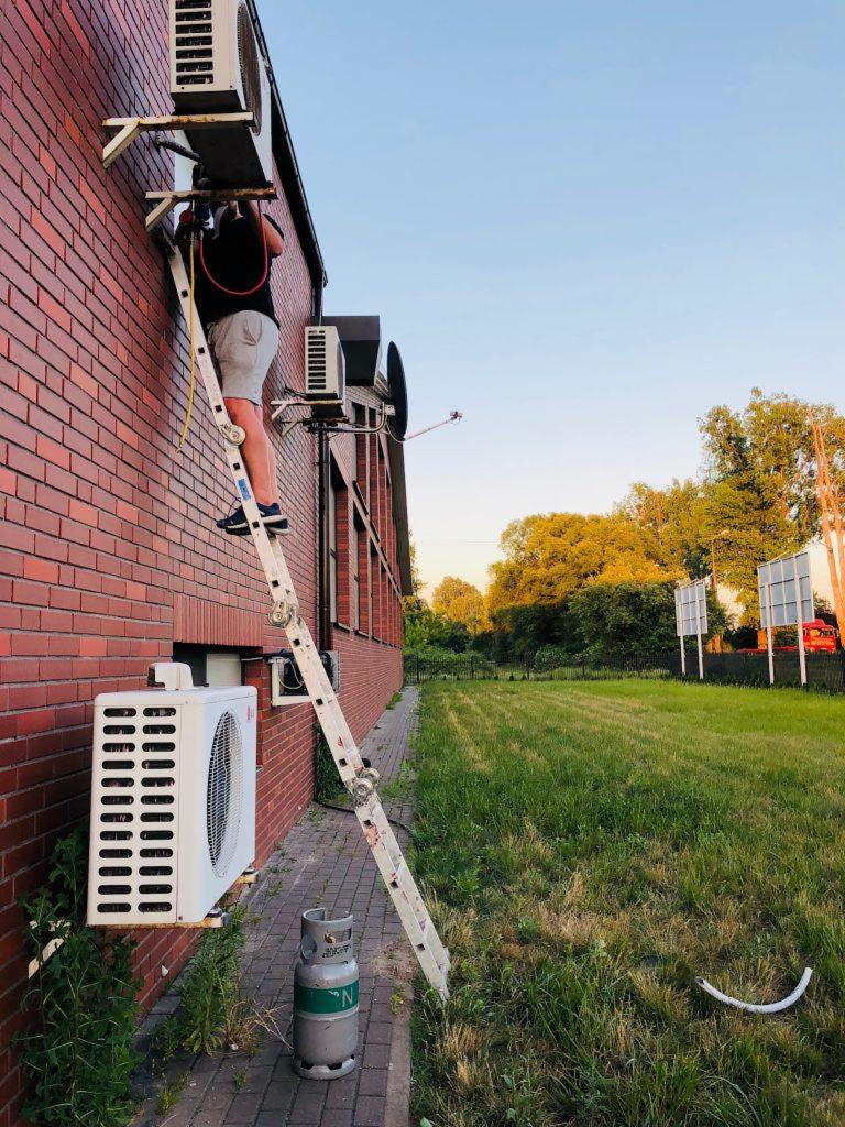 Instalator na drabinie montuje klimatyzator na ceglanej ścianie budynku, butla z gazem stoi obok drabiny, trawa w tle.