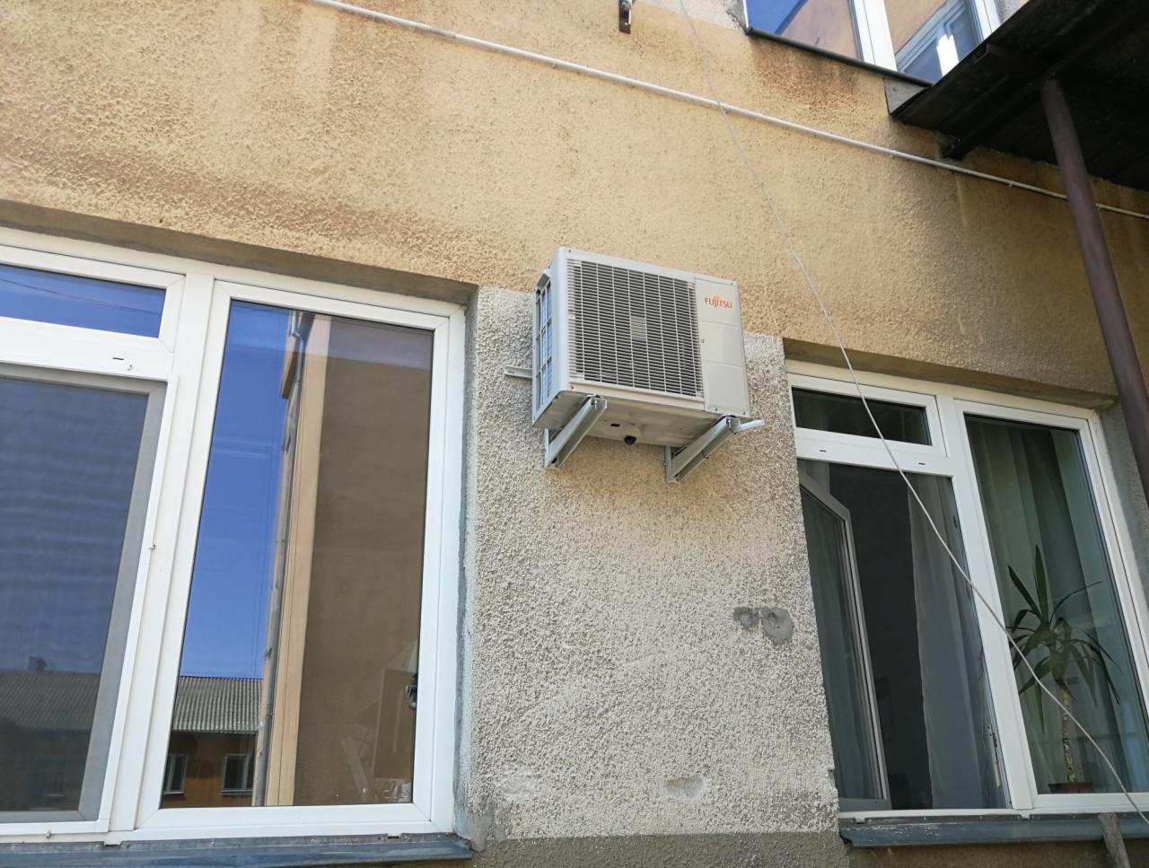 Zamontowana jednostka zewnętrzna klimatyzatora Fujitsu na elewacji budynku w Pruszkowie, widoczna instalacja elektryczna i okno z odbiciem. Budynek o teksturowanej, beżowej elewacji.
