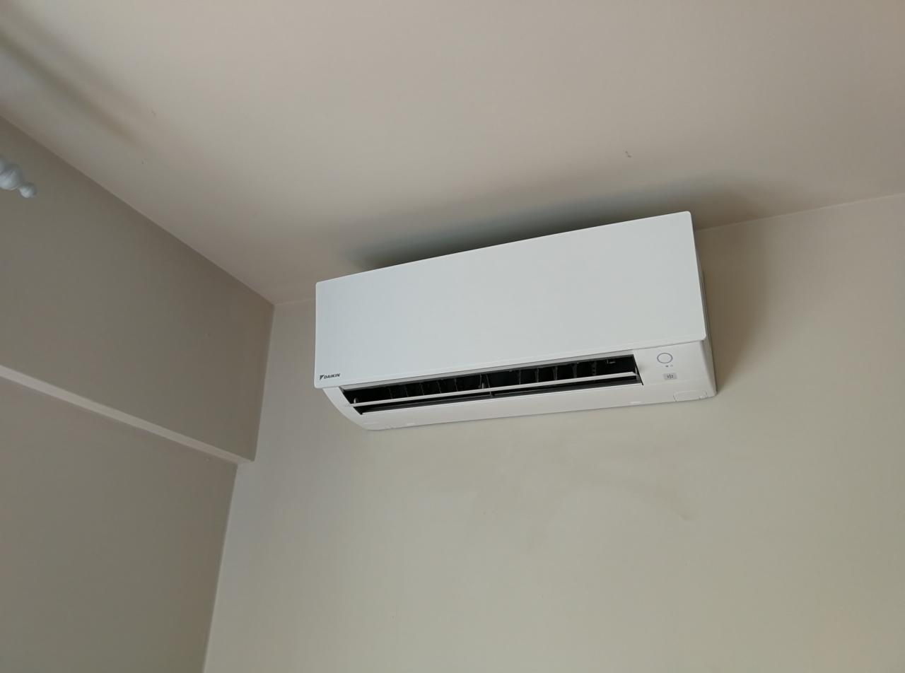 Biała jednostka klimatyzacyjna Daikin zamontowana na beżowej ścianie w rogu pomieszczenia, widoczne detale panelu sterowania i logo producenta.