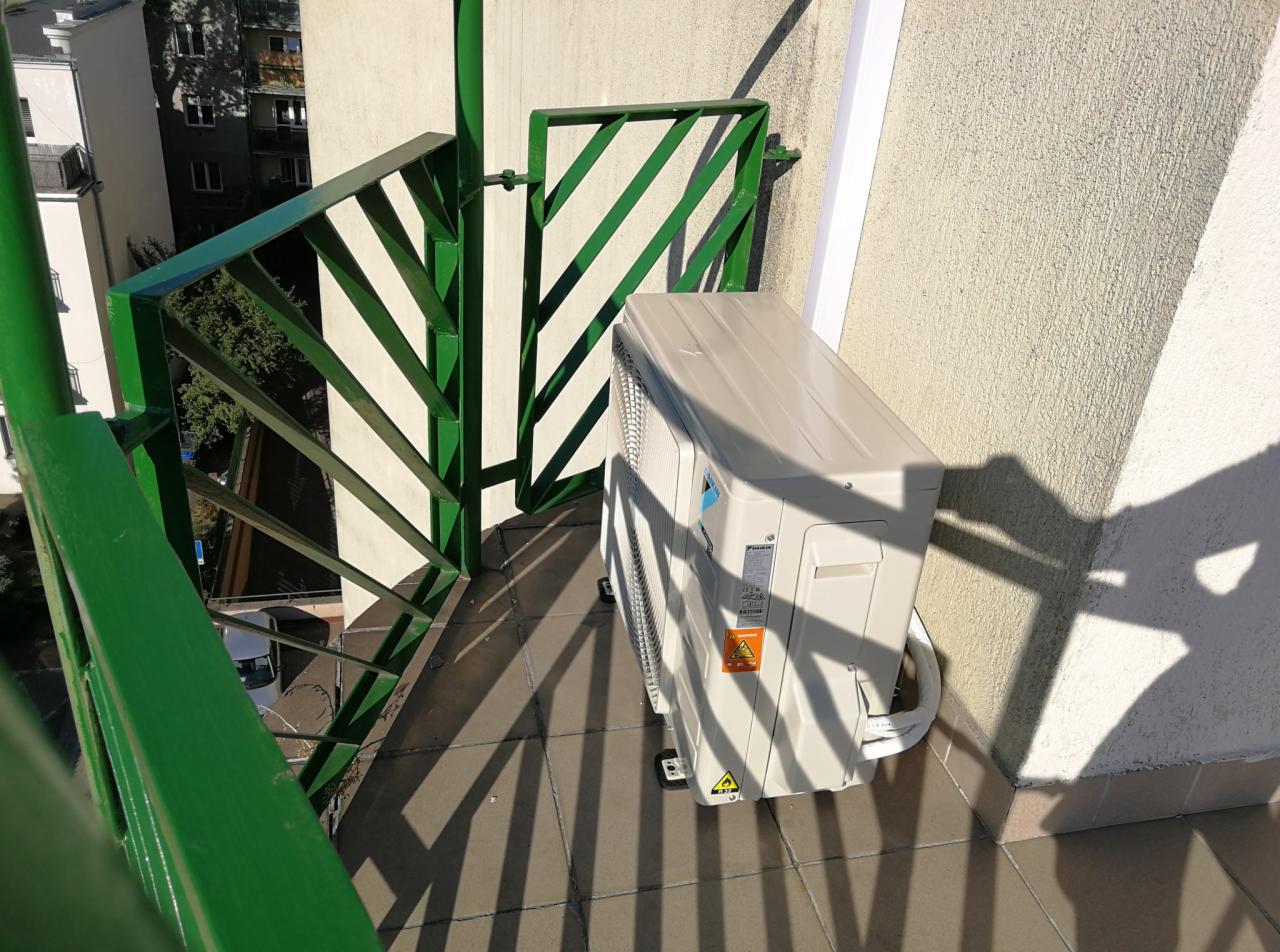 Biała jednostka zewnętrzna klimatyzacji Daikin zamontowana na balkonie z zieloną, ażurową barierką, cień barierki na podłodze.