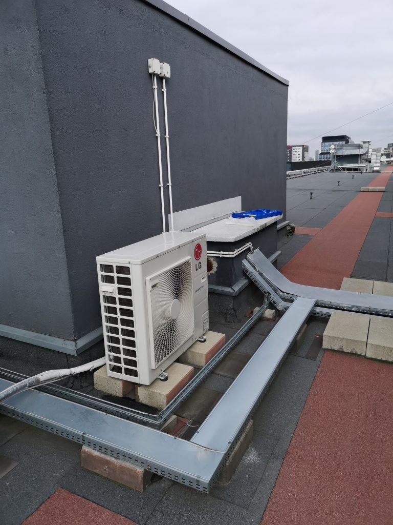 Zewnętrzna jednostka klimatyzacyjna LG umieszczona na ceglanych podstawach na dachu budynku z widocznymi korytami kablowymi i elementami instalacji elektrycznej.