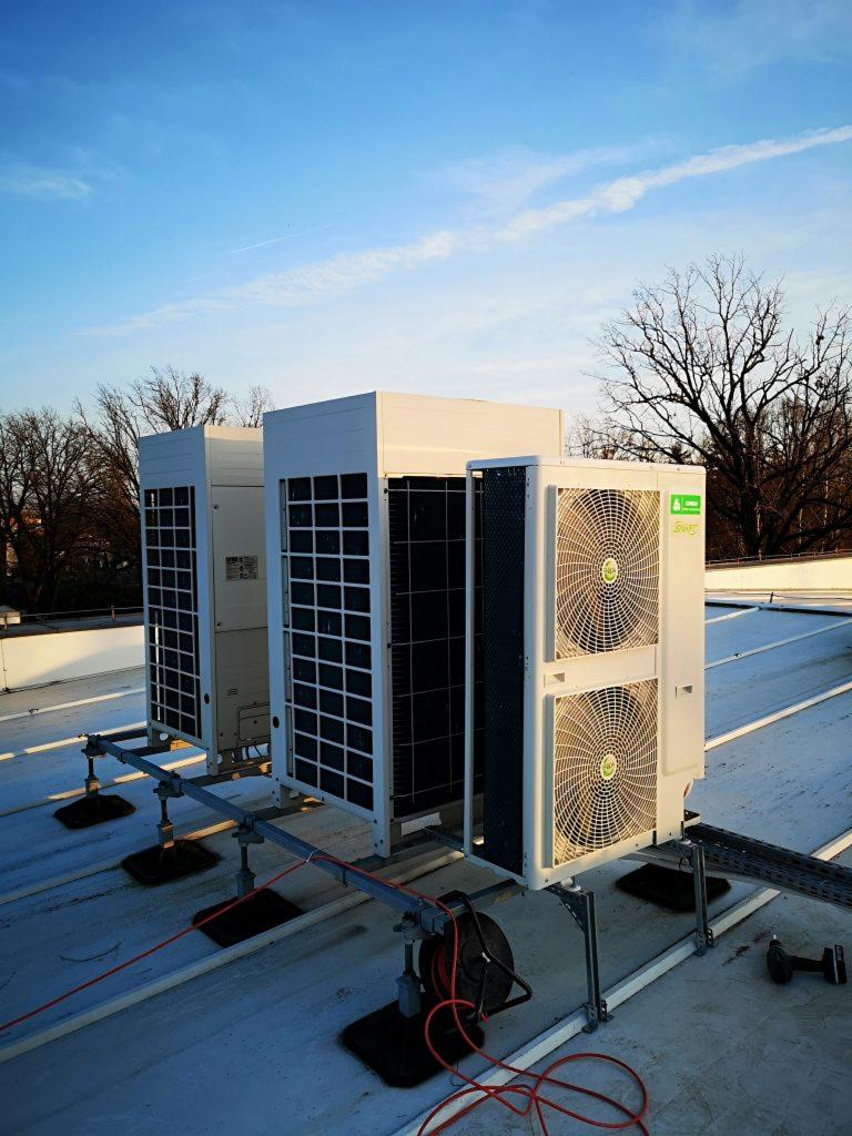 Trzy zewnętrzne jednostki systemu chłodzenia zamontowane na dachu, widoczne przewody elektryczne i narzędzia, błękitne niebo w tle.