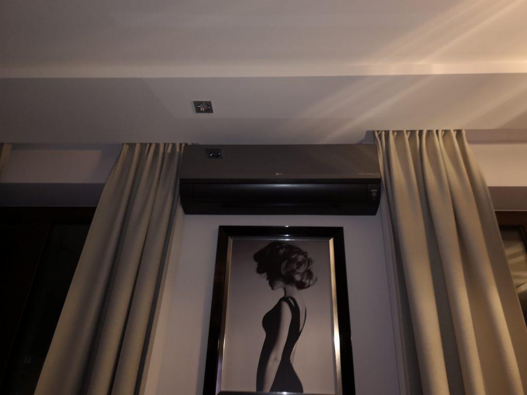 Szara jednostka wewnętrzna zamontowana pod sufitem, obok zasłon i obrazu w czarnej ramie.