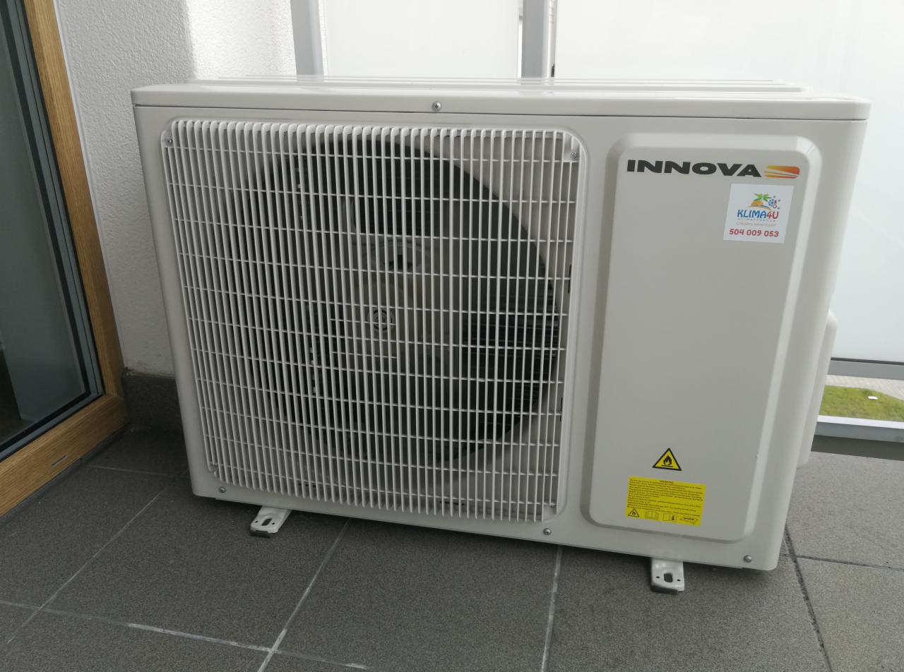 Zewnętrzna jednostka klimatyzatora marki Innova na balkonie, widoczna kratka wentylacyjna, naklejka z logo firmy i numerem telefonu, żółta naklejka ostrzegawcza.