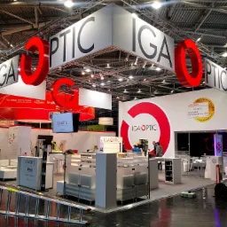 Stoisko targowe IGA OPTIC z dużymi czerwonymi literami 'O' wiszącymi nad przestrzenią wystawienniczą, widoczne elementy konstrukcyjne pod sufitem hali, drabina oparta o podest.