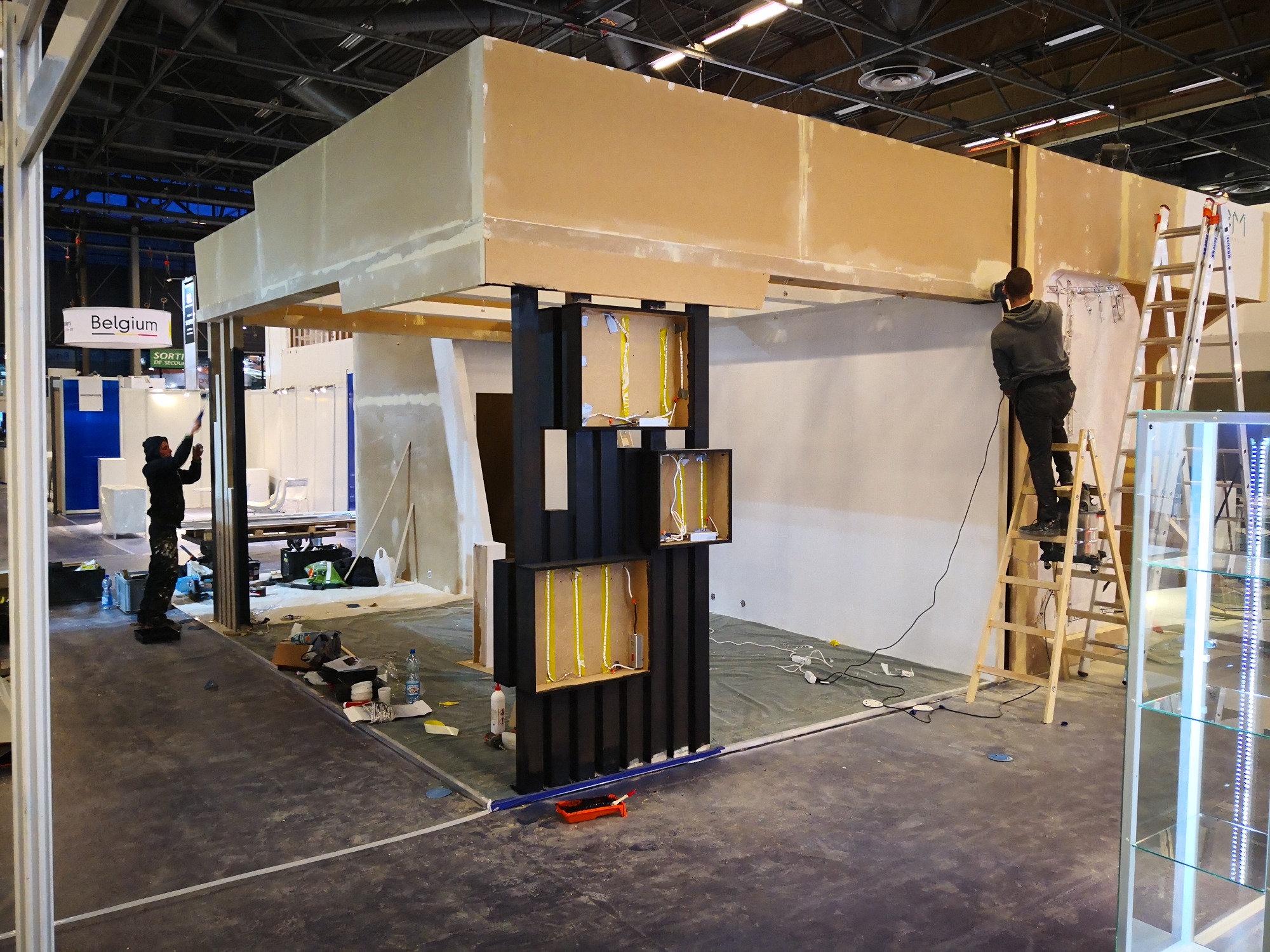 Prace wykończeniowe na stoisku targowym: szpachlowanie sufitu podwieszanego, instalacja oświetlenia LED w czarnej zabudowie, widoczne narzędzia i materiały budowlane na podłodze.