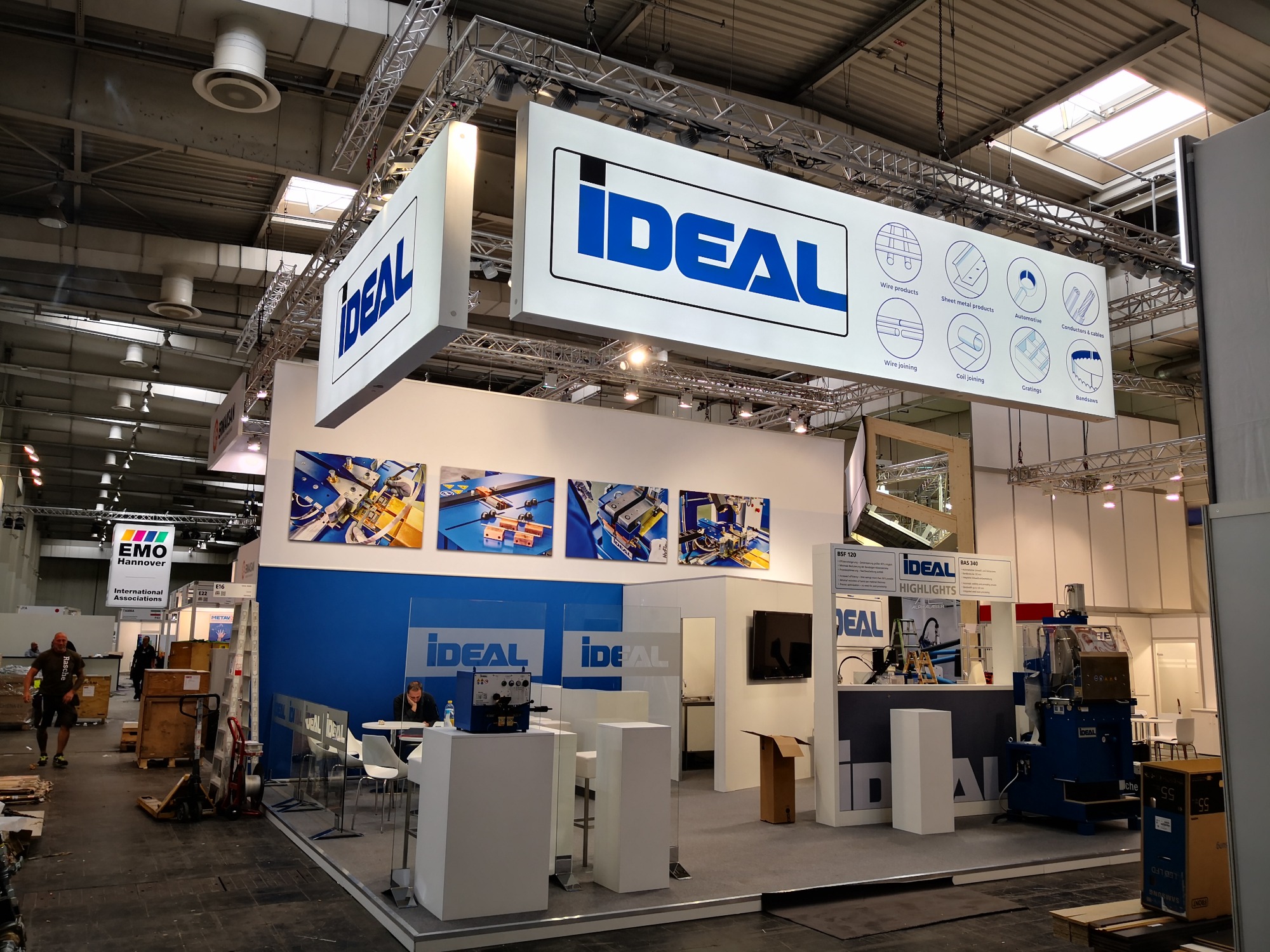 Stoisko targowe firmy Ideal z ekspozycją maszyn do obróbki metali, podwieszane banery informacyjne z logo i opisem produktów, w tle widoczni ludzie i elementy konstrukcji hali.