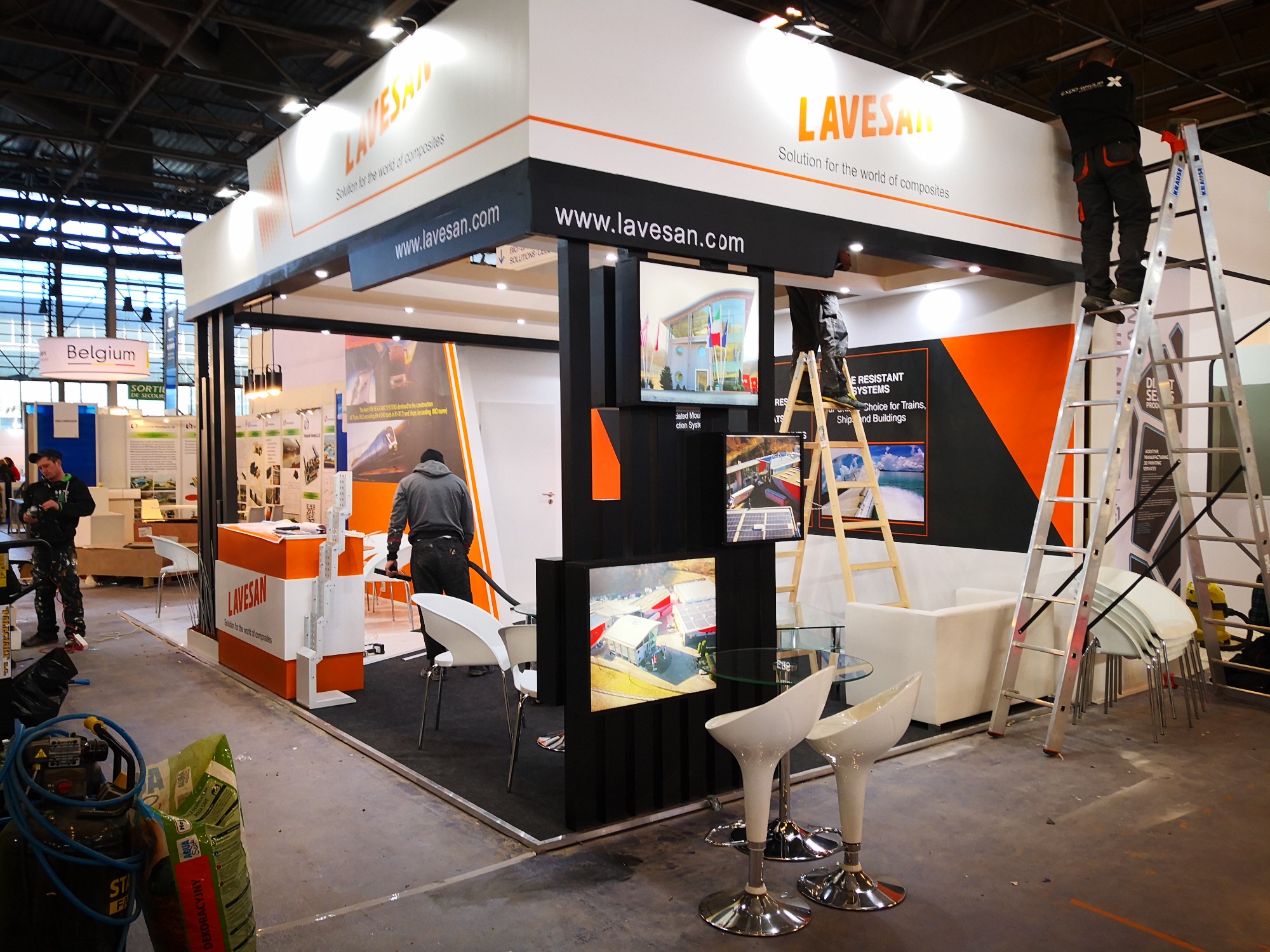 Montaż stoiska targowego Lavesan: dwóch pracowników na drabinach instaluje elementy konstrukcyjne, widoczne monitory z prezentacjami, białe krzesła barowe i sofa, torba z zaprawą dekoracyjną...