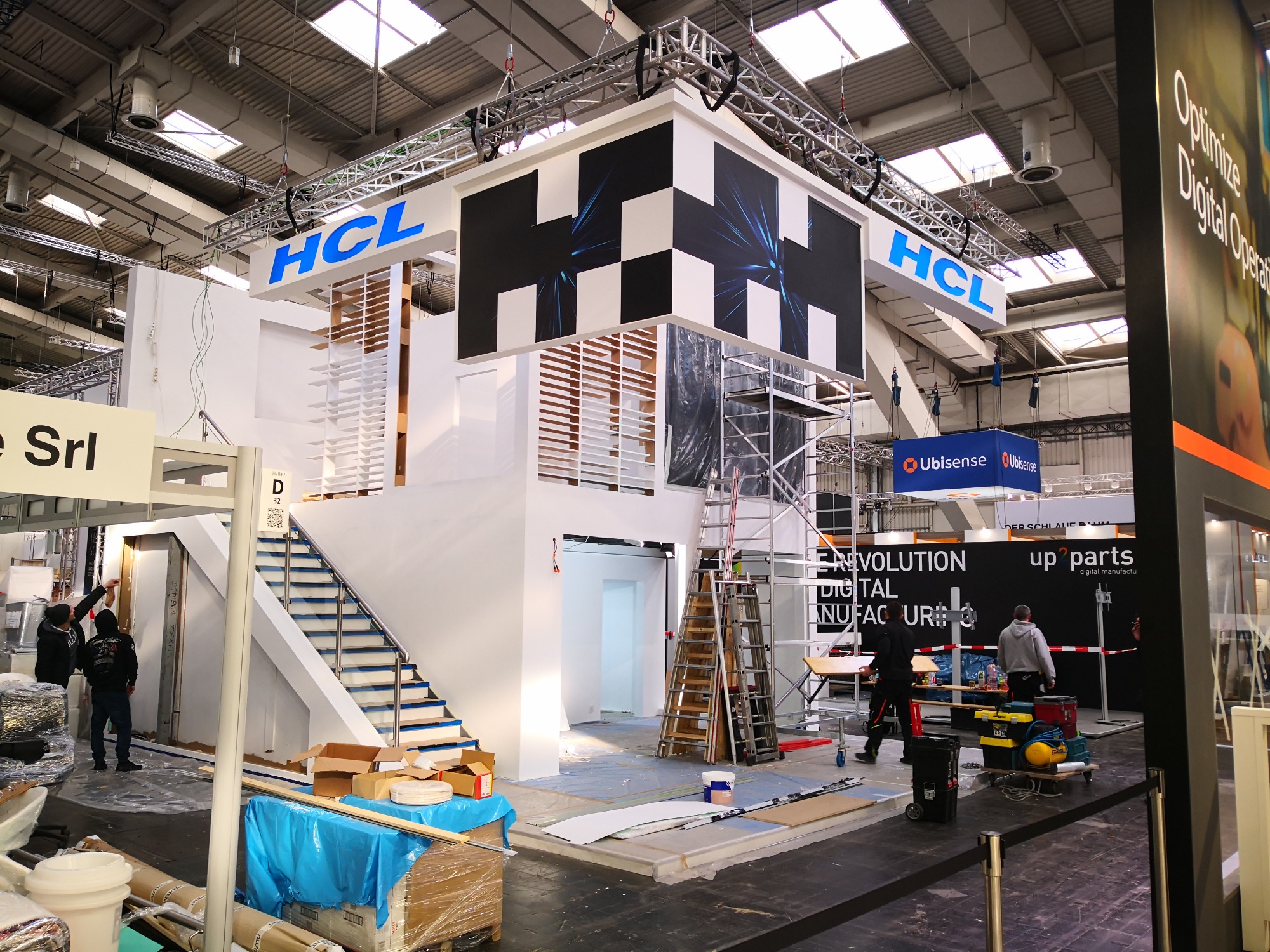 Stoisko targowe w trakcie budowy: widoczne rusztowania, drabiny, materiały budowlane i ekipa montażowa. Konstrukcja z logo HCL i elementami graficznymi w czerni i bieli. Podłoga zabezpieczona folią...